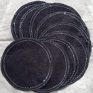May include: A collection of dark denim-colored, circular coasters. Each coaster has a white and light blue zigzag stitched border. The coasters are arranged in a fan shape, showcasing their design and texture.