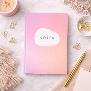 May include: A pink and peach ombre notebook with the word "NOTES" in a white oval. The notebook is surrounded by gold paper clips, a gold pen, a cup of coffee, and small flowers. The text "write your story" is at the bottom of the notebook.