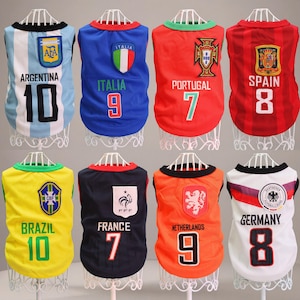 Puede incluir: Ocho camisetas para mascotas, cada una diseñada con los colores y emblemas de diferentes equipos nacionales de fútbol. Las camisetas presentan nombres de equipos, números y logotipos, incluyendo Argentina, Italia, Portugal, España, Brasil, Francia, Países Bajos y Alemania.