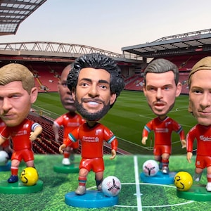 Op de afbeelding: Een verzameling Liverpool FC bobbleheads, elk gekleed in een rood tenue, op een groene basis. De bobbleheads staan op een groene ondergrond met voetballen en een stadion op de achtergrond.