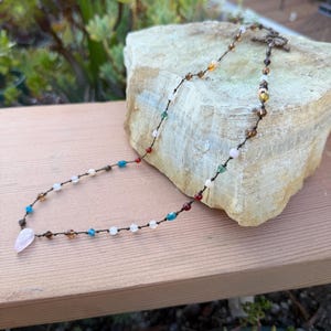 May include: A handmade necklace featuring a variety of colorful beads, including blue, white, brown, and red, strung on a dark cord. The necklace has a unique pendant and a decorative clasp. The necklace is displayed on a light-colored rock.