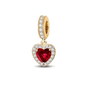 May include: A gold-toned charm featuring a heart-shaped red gemstone at the center, surrounded by a border of small clear stones. The charm includes a cylindrical bail encrusted with clear stones, designed for attachment to a bracelet or necklace.