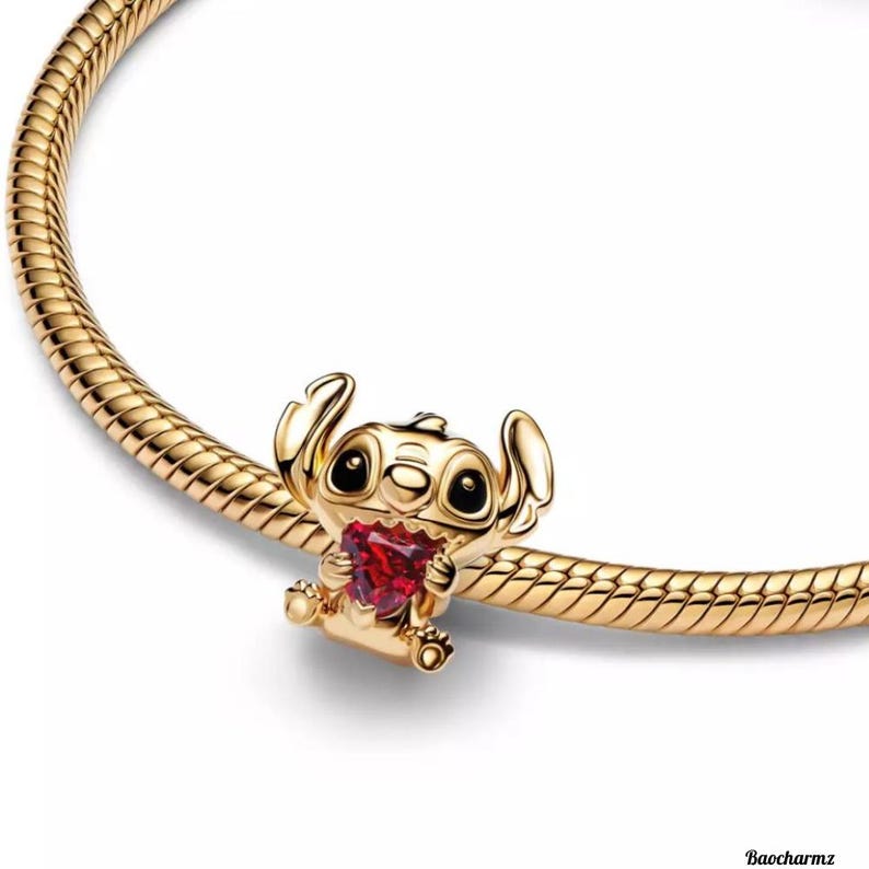 May include: A gold-coloured charm featuring a cartoon character holding a red heart-shaped gem. The charm is attached to a gold-coloured snake chain bracelet. The charm has black eyes and is sitting down.