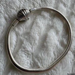 Puede incluir: Una pulsera plateada con un cierre de concha. La pulsera tiene un diseño de cadena de serpiente y se muestra sobre una superficie blanca texturizada. El cierre de concha está detallado con estrías.