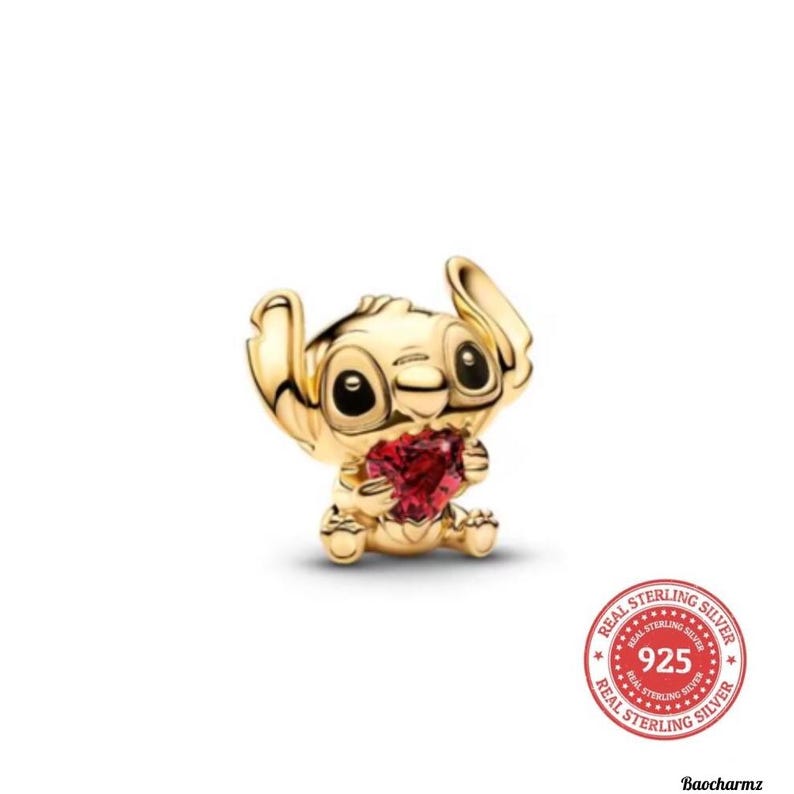 May include: A gold-coloured charm depicting the character Stitch, holding a red heart-shaped gem. The charm has large black eyes and a smiling expression. The image includes a "Real Sterling Silver 925" seal.