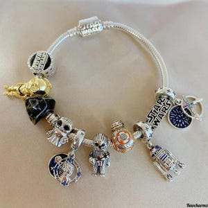 Puede incluir: Una pulsera de plata con abalorios temáticos de Star Wars. Los abalorios representan personajes como R2-D2, Chewbacca y BB-8, junto con logotipos de Star Wars y otras imágenes icónicas. La pulsera se muestra sobre una superficie clara.