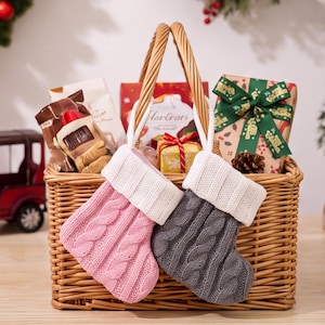 May include: A woven wicker basket containing two small cable-knit Christmas stockings, one light pink and one gray, both featuring white ribbed cuffs. The basket is filled with various gift-wrapped items, including a box with a green ribbon.