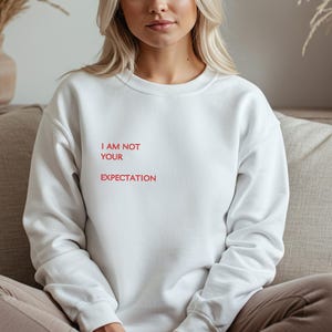 Könnte beinhalten: Weißes Sweatshirt mit dem roten Text "I AM NOT YOUR EXPECTATION". Das Sweatshirt hat einen Rundhalsausschnitt und scheint aus einem weichen Material zu sein. Der Text ist mittig auf der Brust platziert.