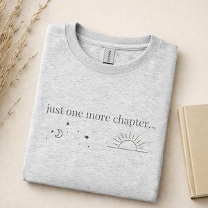 Könnte beinhalten: Hellgraues Rundhals-T-Shirt mit dem Text "just one more chapter..." und einer Grafik aus Mond, Sternen und Sonne. Das T-Shirt ist sauber gefaltet.