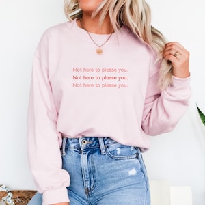 Könnte beinhalten: Hellrosa Sweatshirt mit dem Text "Not here to please you" in wiederholtem Muster. Dazu eine blaue Jeans. Der Look wirkt lässig und bequem.
