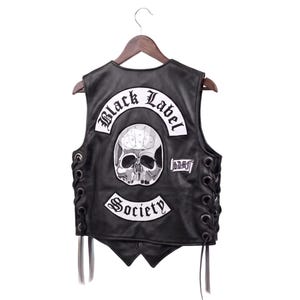 May include: A black leather waistcoat featuring a large white patch on the back with the text Black Label Society and a skull graphic. The sides of the waistcoat are laced with black leather cords, and a small rectangular patch with text is on the right side.