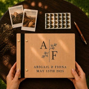 Könnte beinhalten: Ein rechteckiges Fotoalbum aus Holz mit schwarzem Buchrücken und goldenen Metallschrauben. Der Einband zeigt die eingravierten Buchstaben A und F, getrennt durch eine vertikale Linie mit Blattmotiven, sowie den Text ABIGAIL & FIONA MAY 15TH 2025.