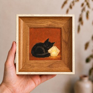 May include: A square framed painting featuring a black cat curled up and sleeping next to a glowing, geometric, white paper lantern. The artwork is set against a textured, deep orange background and encased in a light-toned wooden frame.