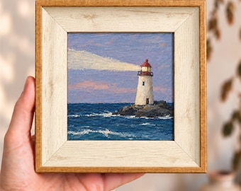 May include: A square oil painting of a white lighthouse with a red top, situated on a dark rocky outcrop amidst a deep blue sea. A bright beam of light extends from the lighthouse into a purple and pink sky, all encased in a light wood frame.