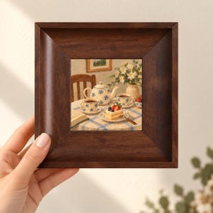 May include: A square wooden frame containing a painting of a tea set on a blue and white chequered tablecloth. The scene depicts a teapot, two teacups, a slice of cake topped with strawberries and blueberries, and a vase of white flowers.