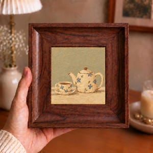 Peut inclure: Une peinture à l'huile carrée dans un cadre en bois brun foncé représentant une théière crème et une tasse assortie avec des motifs floraux bleus. L'œuvre présente un fond vert clair et une surface beige, illustrant une nature morte de thé.