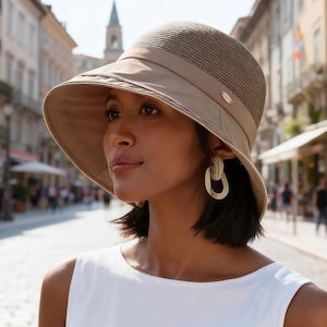 Può includere: Un cappello da sole beige con tesa larga e corona strutturata. Il cappello ha una fascia intorno alla corona e un piccolo dettaglio dorato. La donna indossa un top bianco e orecchini dorati.