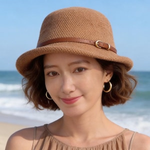 May include: A brown cloche hat with a brown leather band and gold buckle. The hat has a rounded crown and a slightly wider brim. The background shows a beach and the sea.