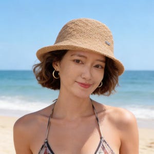 May include: A woman wearing a tan, woven sun hat with a small metal tag. The hat has a rounded crown and a slightly wider brim. She is also wearing gold hoop earrings and a patterned halter top. The background shows a beach and ocean.
