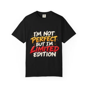 Può includere: Una t-shirt nera a maniche corte con il testo I'M NOT PERFECT BUT I'M LIMITED EDITION stampato in un carattere in stile pennellata. Le parole sono colorate in bianco, giallo e rosso, centrate sulla parte anteriore del capo.