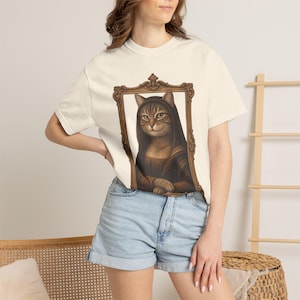 May include: A cream-colored short-sleeve t-shirt featuring a graphic print of a brown tabby cat wearing a dark hood, positioned inside an ornate gold-colored rectangular frame. The cat is depicted with its paws crossed in a classic portrait style.