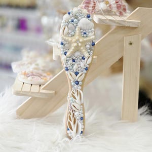 Op de afbeelding: Houten haarborstel versierd met schelpen, parels en een zeester. De borstel heeft een licht houten handvat en is versierd met witte, blauwe en crèmekleurige versieringen. Het ontwerp roept een kust- of nautisch thema op.