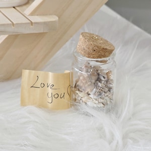 Puede incluir: Un pequeño tarro de cristal lleno de conchas marinas, coronado con un tapón de corcho. Una nota manuscrita que dice "Love you" está adjunta al tarro. El tarro está sobre una superficie blanca y esponjosa.