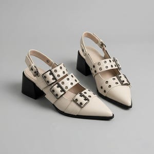 May include: A pair of cream-colored slingback heels with pointed toes and black block heels. The shoes feature multiple straps with silver-tone buckles and eyelet details, offering a modern, edgy aesthetic.