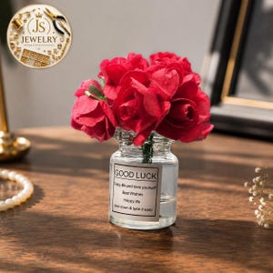 Puede incluir: Un pequeño frasco de vidrio transparente lleno de rosas artificiales de color rojo vibrante. El frasco presenta una etiqueta rectangular blanca con el texto en negro: GOOD LUCK, Enjoy life and love yourself, Best Wishes, Happy life, Slow down & take it easy.