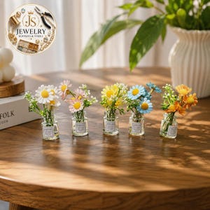 May include: Five miniature glass bottles, each containing a small bouquet of artificial daisy-like flowers in white, pink, yellow, blue, and orange. Each clear bottle features a small square label with the text Good Luck and Pretty Flower.
