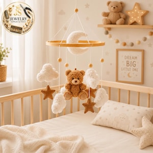 May include: A baby mobile featuring a central brown teddy bear, a white crescent moon, and several white clouds and brown stars suspended from a circular wooden frame by white strings. The mobile is designed for nursery decor.