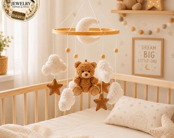 May include: A baby mobile featuring a central brown plush teddy bear, a white crescent moon, and several white fluffy clouds and brown felt stars suspended from a circular wooden frame by white strings with small wooden beads.