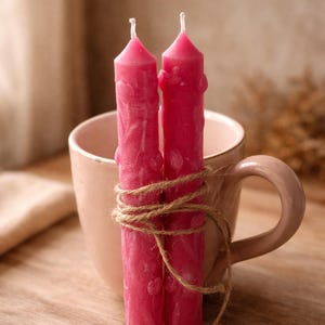 Puede incluir: Dos velas de color rosa brillante, atadas con hilo, se colocan frente a una taza rosa pálido. Las velas tienen una superficie texturizada y un diseño sencillo. La taza tiene un asa curvada y un acabado liso.