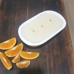 Puede incluir: Una vela blanca de forma ovalada con tres mechas, colocada en un recipiente blanco. Rodajas de naranja están dispuestas alrededor de la vela sobre una superficie de madera oscura. La vela es de color crema blanquecino.