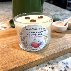 Puede incluir: Una vela de vidrio con tres mechas de madera y cera de color crema. La vela tiene una etiqueta con el texto "It's a beautiful day to save lives (but I'd rather be napping)". La vela está sobre una tabla de cortar de madera.