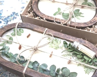 May include: Two oval-shaped candles in wooden trays, decorated with green foliage and tied with twine. The candles are in a cardboard box. The text "NEW GREEN FOLIAGE CANDLES!" and "Handcrafted and beautiful!" are visible.