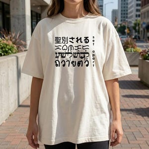 Puede incluir: Camiseta color crema con texto negro en varios idiomas. El texto incluye caracteres japoneses, hebreos y tailandeses, junto con las palabras en inglés "set apart" verticalmente a la derecha. La camiseta tiene mangas cortas y un corte holgado.