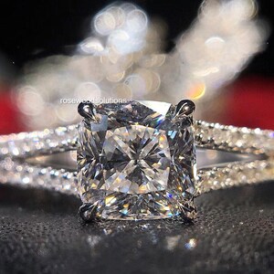 May include: A diamond ring featuring a large, square-cut central stone. The split band is adorned with smaller diamonds, enhancing the sparkle. The ring is set against a dark backdrop, accentuating the brilliance of the gemstones.