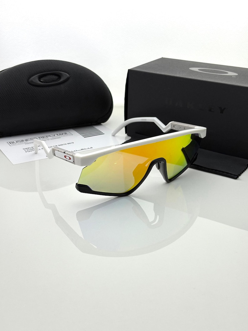 K&ouml;nnte beinhalten: Wei&szlig; gerahmte Sonnenbrille mit gelb-orange verspiegelten Gl&auml;sern. Die Sonnenbrille wird mit einem schwarzen Etui und einer schwarzen Schachtel mit dem Markennamen pr&auml;sentiert. Ein Reinigungstuch und Papiere sind ebenfalls sichtbar.