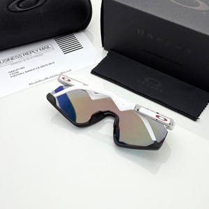 K&ouml;nnte beinhalten: Wei&szlig;e und schwarze Sonnenbrille mit verspiegelten Gl&auml;sern, einem schwarzen Etui und einer schwarzen Box. Die Sonnenbrille hat ein modernes Design mit wei&szlig;em Rahmen und roten Akzenten. Auf der Box steht der Markenname "OAKLEY".