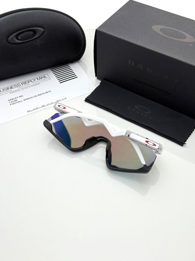 K&ouml;nnte beinhalten: Wei&szlig;e und schwarze Sonnenbrille mit verspiegelten Gl&auml;sern, einem schwarzen Etui und einer schwarzen Box. Die Sonnenbrille hat ein modernes Design mit wei&szlig;em Rahmen und roten Akzenten. Auf der Box steht der Markenname "OAKLEY".