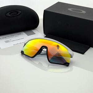 K&ouml;nnte beinhalten: Wei&szlig;e und schwarze Sportsonnenbrille mit verspiegelten orangefarbenen und gelben Gl&auml;sern. Die Sonnenbrille liegt neben einem schwarzen Etui, einer schwarzen Box, einem Reinigungstuch und einem Gesch&auml;ftssendungsdokument.