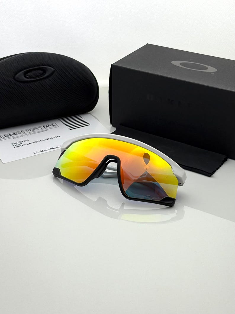 K&ouml;nnte beinhalten: Wei&szlig;e und schwarze Sportsonnenbrille mit verspiegelten orangefarbenen und gelben Gl&auml;sern. Die Sonnenbrille liegt neben einem schwarzen Etui, einer schwarzen Box, einem Reinigungstuch und einem Gesch&auml;ftssendungsdokument.