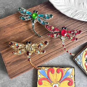 Pode incluir: Três broches em forma de libélula com corpos metálicos e asas incrustadas com strass facetados. Um broche é verde, um é vermelho e um é dourado, cada um com uma cauda longa segmentada e detalhes intrincados de cristal.