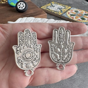 May include: Two silver-colored Hamsa hand charms. One features an eye design, the other has fish and floral details. Both have a loop at the bottom for attaching to jewelry or other crafts. The charms are detailed with intricate patterns.