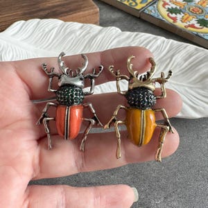 Puede incluir: Dos broches decorativos de escarabajo. Uno tiene un cuerpo naranja y detalles de gemas verdes, el otro tiene un cuerpo amarillo y detalles de gemas negras. Ambos tienen detalles metálicos plateados y dorados, incluyendo antenas y patas.