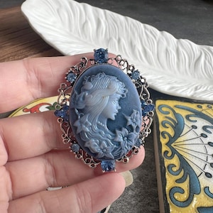 May include: An oval brooch featuring a blue cameo of a profile portrait of a person with long hair and flowers. The cameo is set in a silver-toned filigree metal frame decorated with small, round, faceted blue crystals along the outer edge.