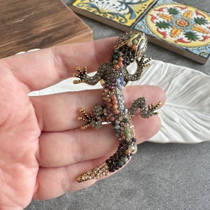 Op de afbeelding: Een gedetailleerde hagedisbroche, versierd met een verscheidenheid aan kleurrijke edelstenen. De broche heeft een gedetailleerd ontwerp met goudkleurige accenten. De edelstenen zijn in bruin-, groen-, oranje- en paarstinten.