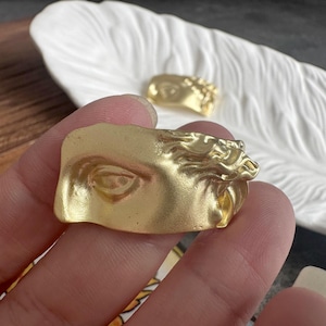 Può includere: Un anello color oro caratterizzato da un rilievo scolpito raffigurante un occhio umano e capelli fluenti. Il metallo presenta una finitura opaca con contorni dettagliati che imitano tratti artistici classici. La fascia è larga e di forma irregolare.