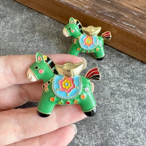 Op de afbeelding: Twee groene paardvormige emaille broches met goudkleurige accenten, een lichtblauw zadel met een geel en roze bloemmotief, en zwart-rode staartdetails. Elke broche heeft een goudkleurige baarvorm en een kleine heldere kristal.
